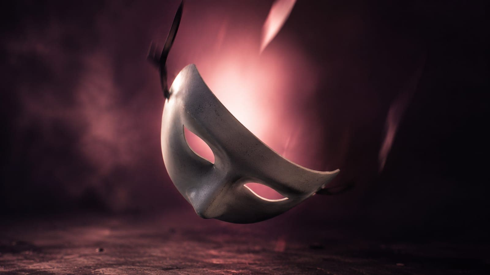 A theatrical mask falling away from a darkened studio backdrop, revealing soft pink light behind it