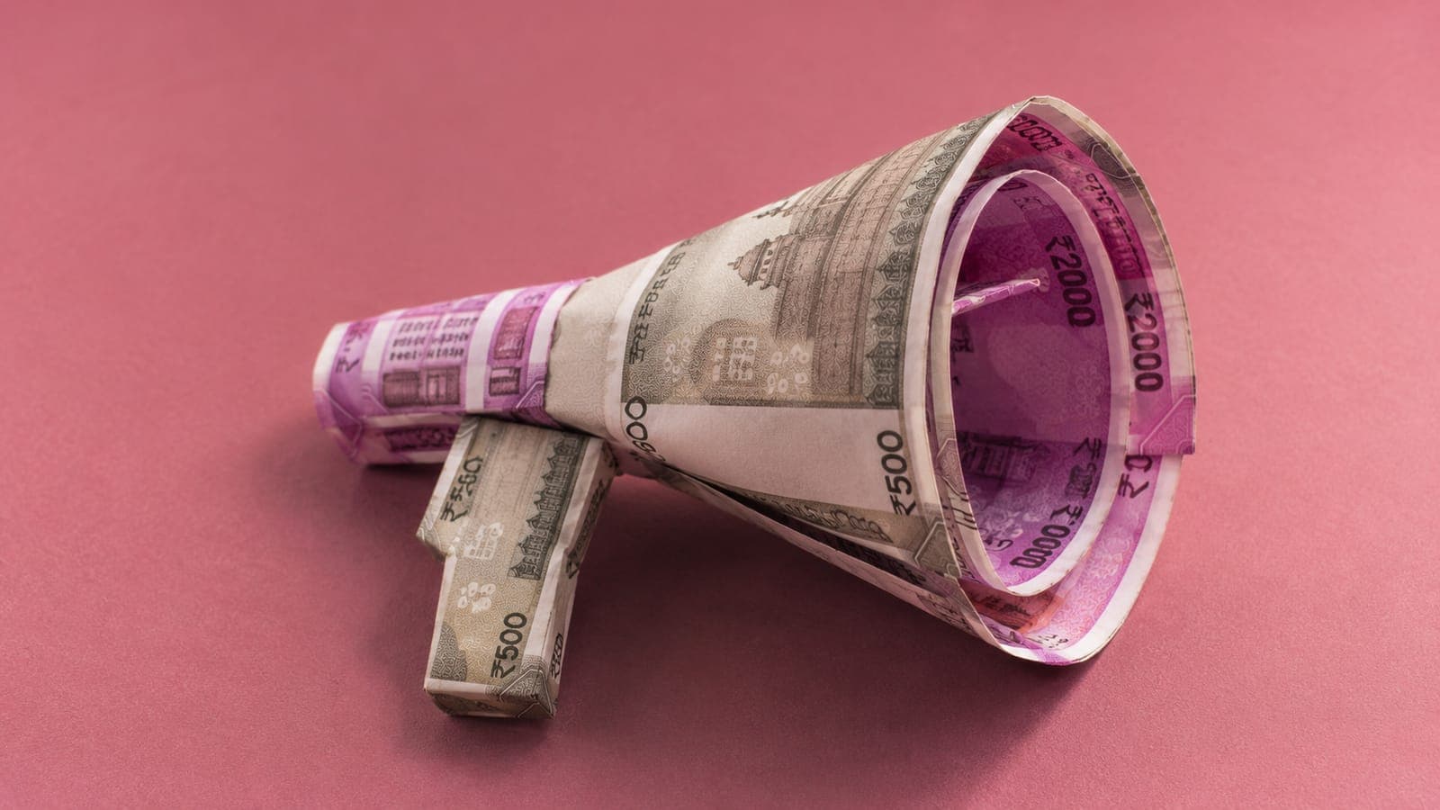 Indian rupee notes folded into a megaphone shape on a soft pink studio backdrop