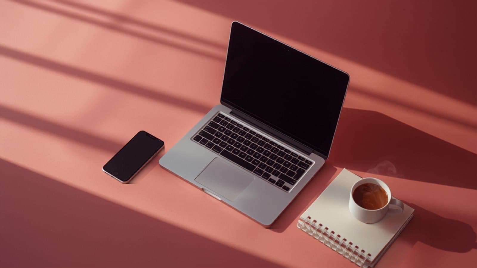 A laptop open on a warm pink desk beside a phone, a spiral notebook and a cup of chai, illustrating an Indian creator writing a pitch email to a brand